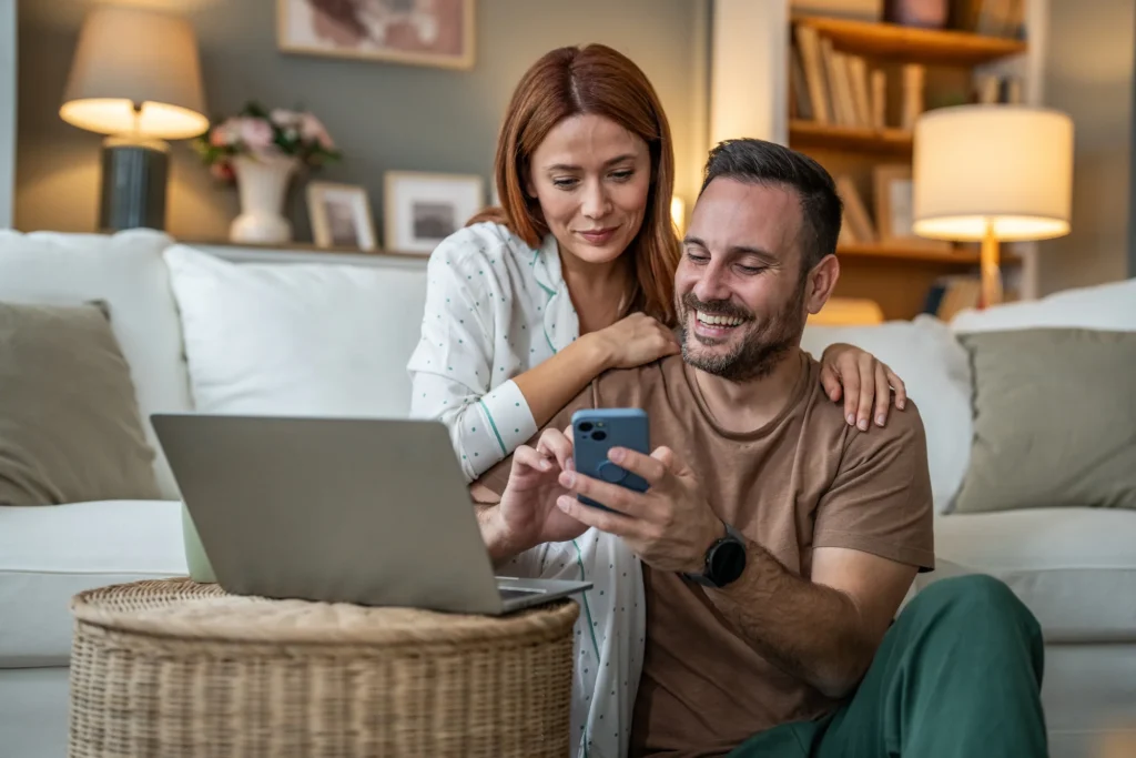 Couple smiling at smartphone and laptop at home
